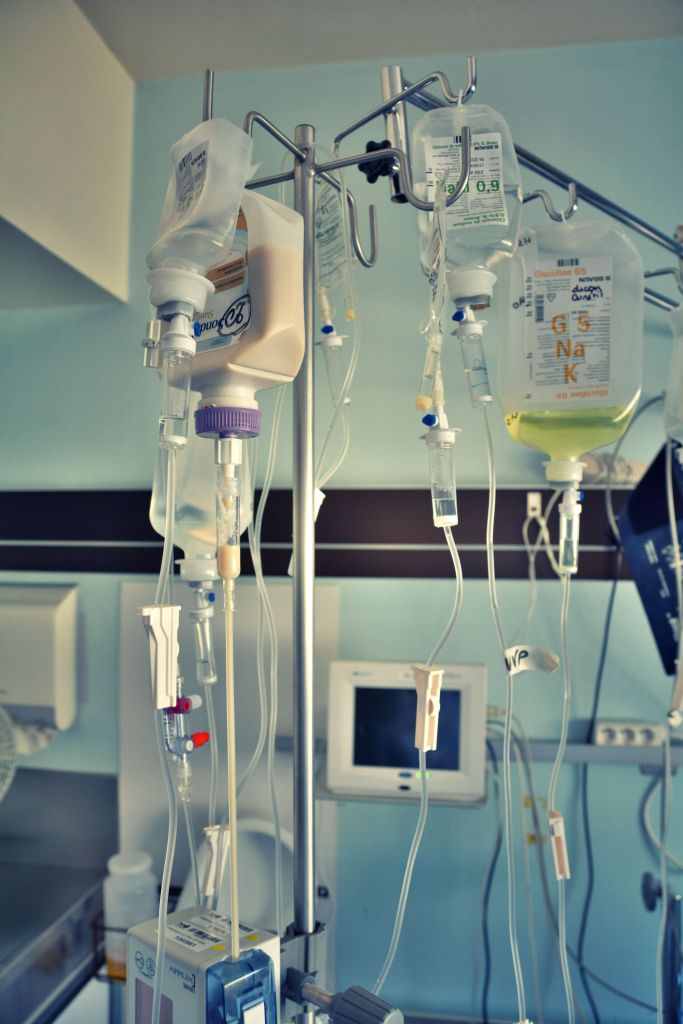 Photo shows a medical stand in what seems to be a hospital room, with several bags hanging upside down and tubes going down. One of the bags is bigger and has a cream like content.