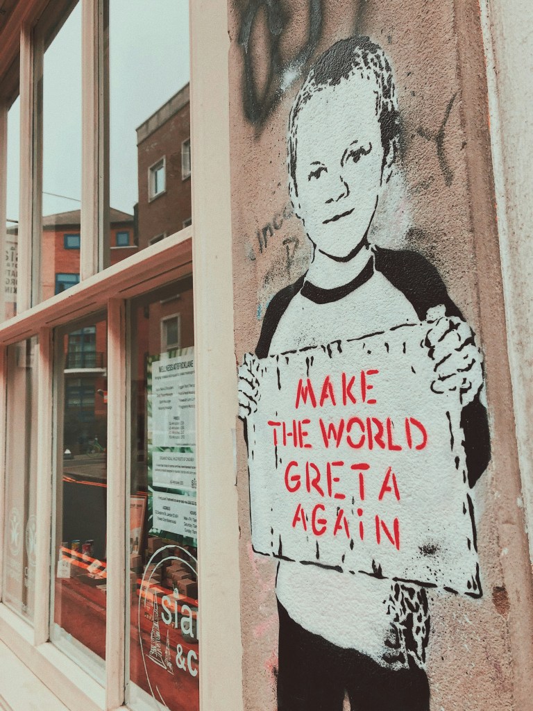 Photo of graffitti on wall. Image of a child next to windows, child is holding a sign that reads: Make the World Greta again. 