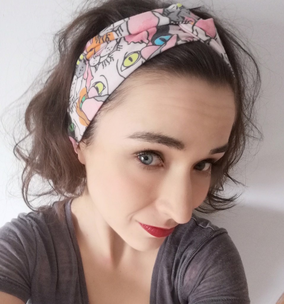 Close up selfie of Alicia looking into the lens, wearing a head band, semi long brown hair backwards, red lipstick, brown T-shirt.