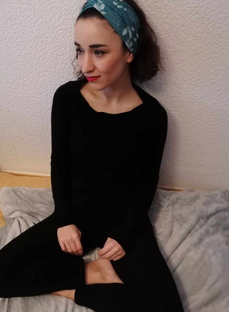Alicia wearing all black (trousers and jumper) sitting cross legged on a grey blanket against a white wall, looking sideways away from camera