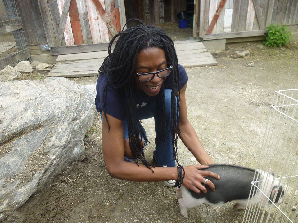Christopher Sebastian wearing blue jeans and navy blue T-shirt, kneeling down on one leg, holding hand on the back of a small pig, a shed in the background