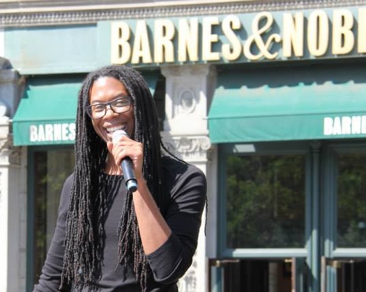 Christopher Sebastian, long dreads  handing mic in left hand, smiling, back ground is shop with sign 'barnes & Nobles'. 