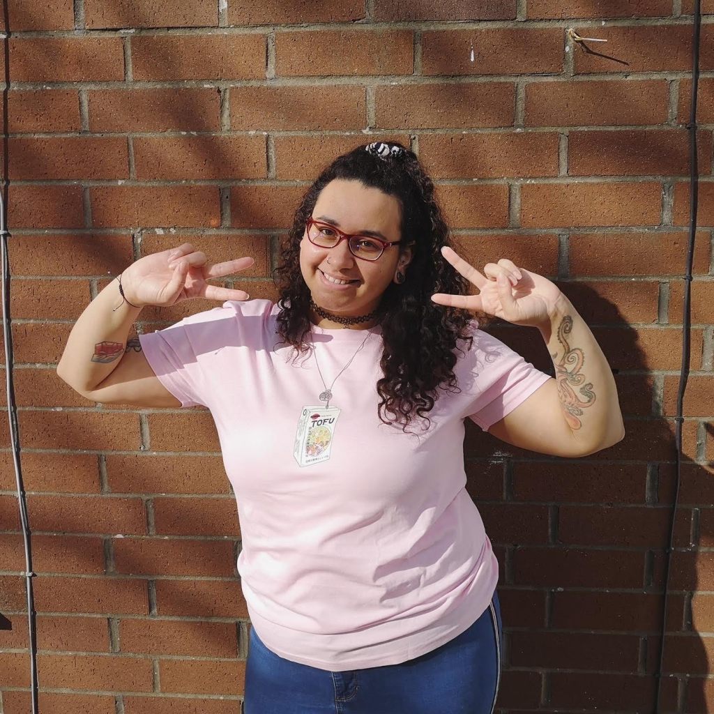 Jade is standing with back against a red brick wall, wearing pink T-shirt and jeans, smiling, both arms raised sidewards and elbows bent with hand making V-sign pointed towards face.  