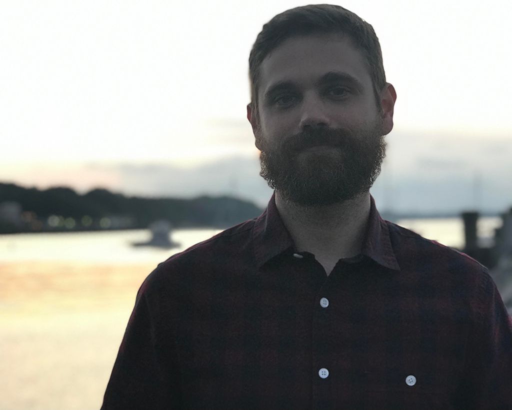 photo of person wearing dark shirt, dark beard and moustache, bright background (appearing to be a sea / lake)  