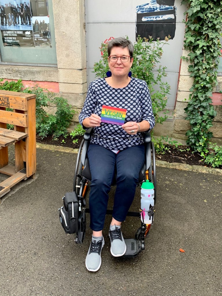 Person with short dark/grey hair, glasses, wearing blue/white dotted shirt and blue trousers, sitting in black/orange wheelchair, wall with some foliage in the back. Geertrui is holding a postcard reading 'VEGAN' against a background of the rainbow flag. 
