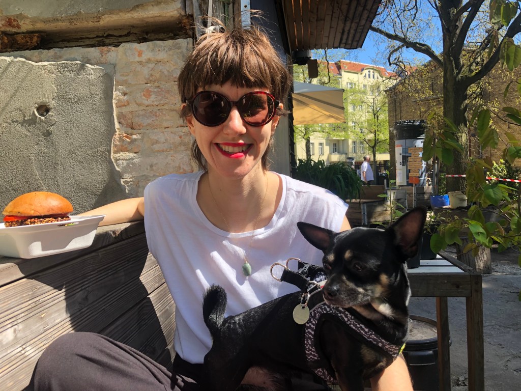 Photo of person wearing sunglasses, white T-shirt, sitting on an outdoors terrace, small black dog on lap, burger in take away dish on the side. 