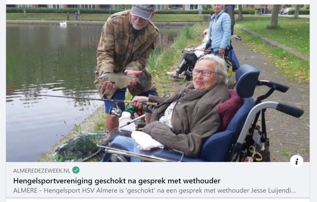 [ID: persons at side of a lake with fishing gear, one standing holding a fish in both hands, posing at camera, other people sitting down looking at camera]
