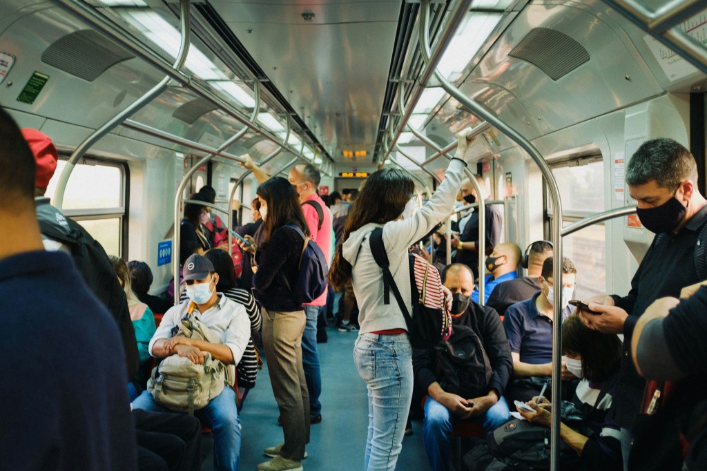 Photo of a subway carriage with all seats taken, and several people standing up. People are wearing face masks. 